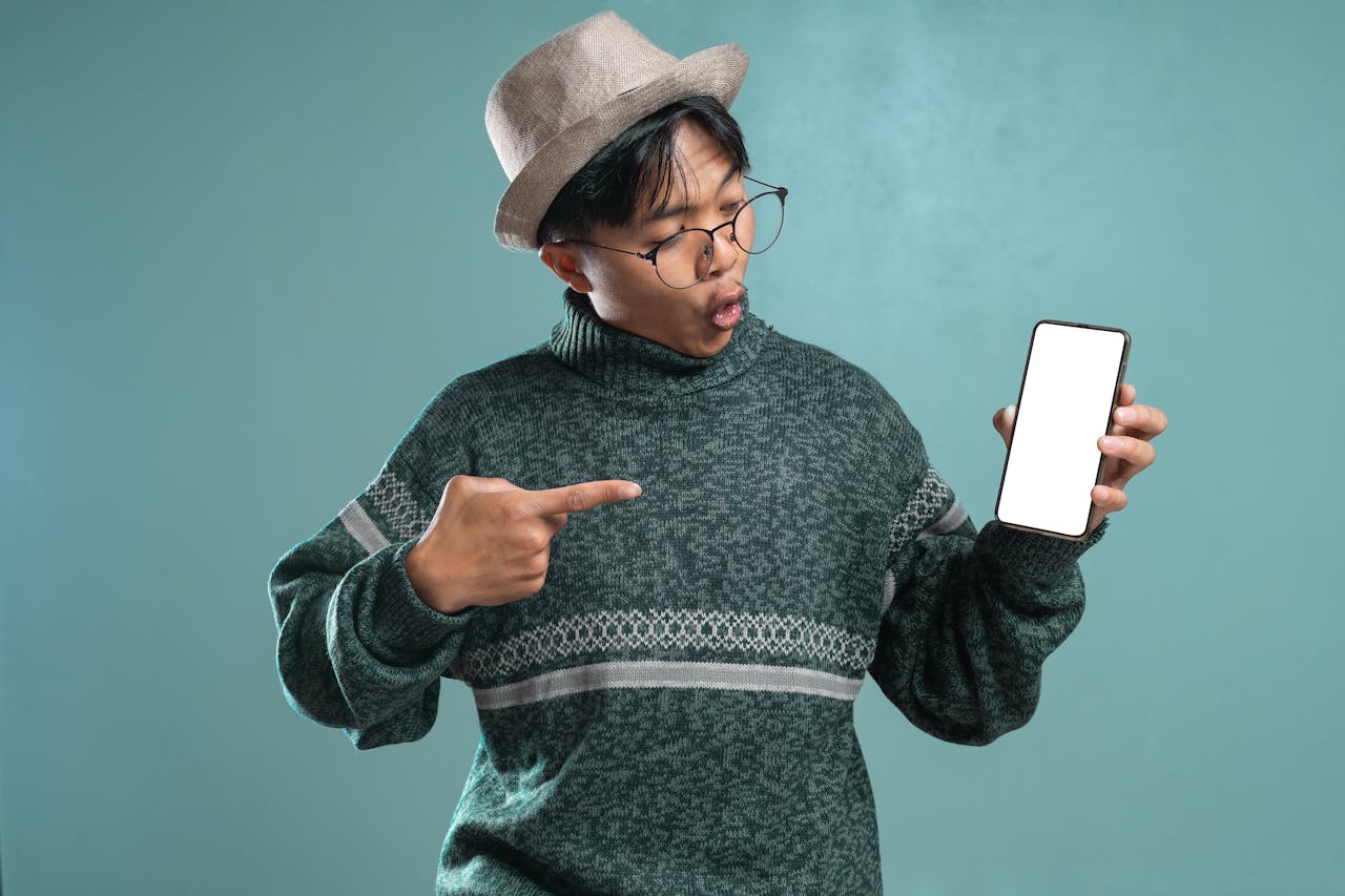 Young man in green sweater and hat pointing at blank smartphone screen with excitement.